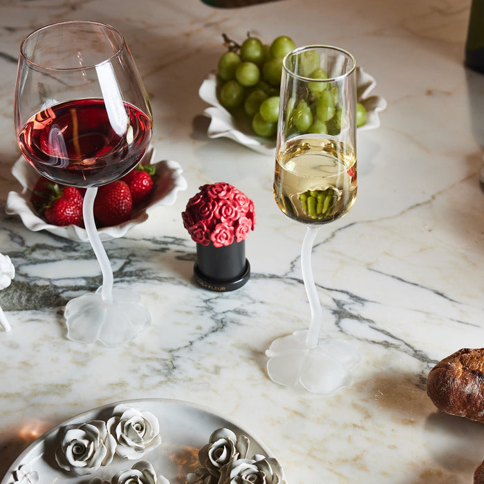 Two uniquely designed wine glasses with twisted white stems on a marble surface; one glass contains red wine with strawberries behind it, the other contains white wine with green grapes behind it, surrounded by decorative rose-shaped objects and a piece of bread.