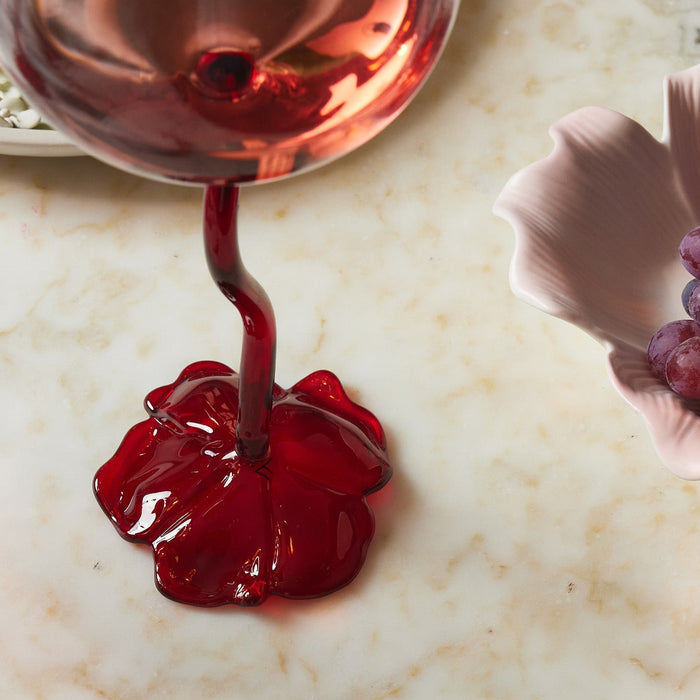 Close-up of a red glass wine glass stem with a flower-shaped base on a marble surface next to a pink dish with grapes.