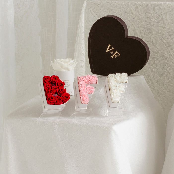 Flower arrangements shaped as letters B, F, and F with red, pink, and white roses on a white tablecloth, next to a dark heart-shaped box with the text 'VeF'.