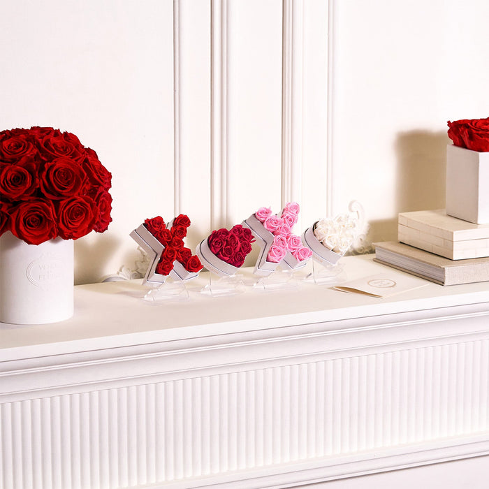 White mantel with red and pink rose arrangements shaped as X and O letters, a large bouquet of red roses in a white Venus Et Fleur vase, and stacked books on the right.