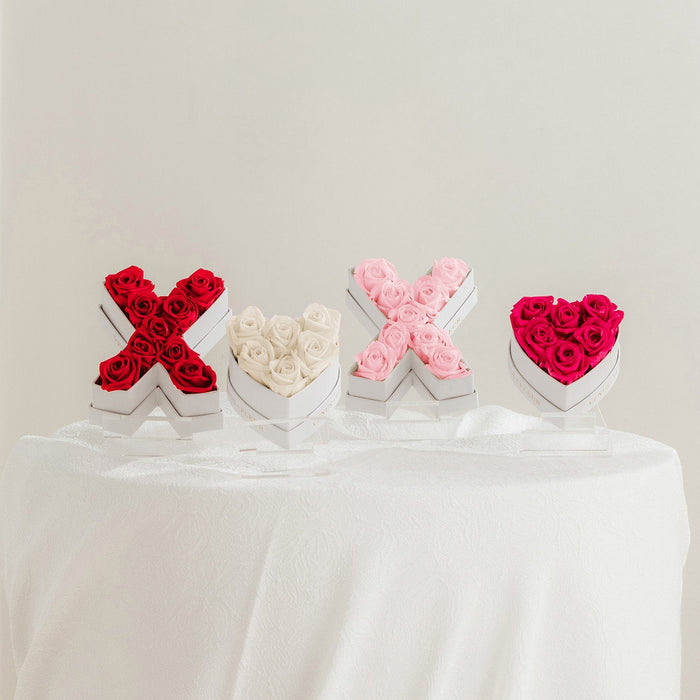 Four floral arrangements on a white tablecloth, shaped as X and heart symbols: red roses in an X shape, white roses in a heart shape, pink roses in an X shape, and red roses in a heart shape.